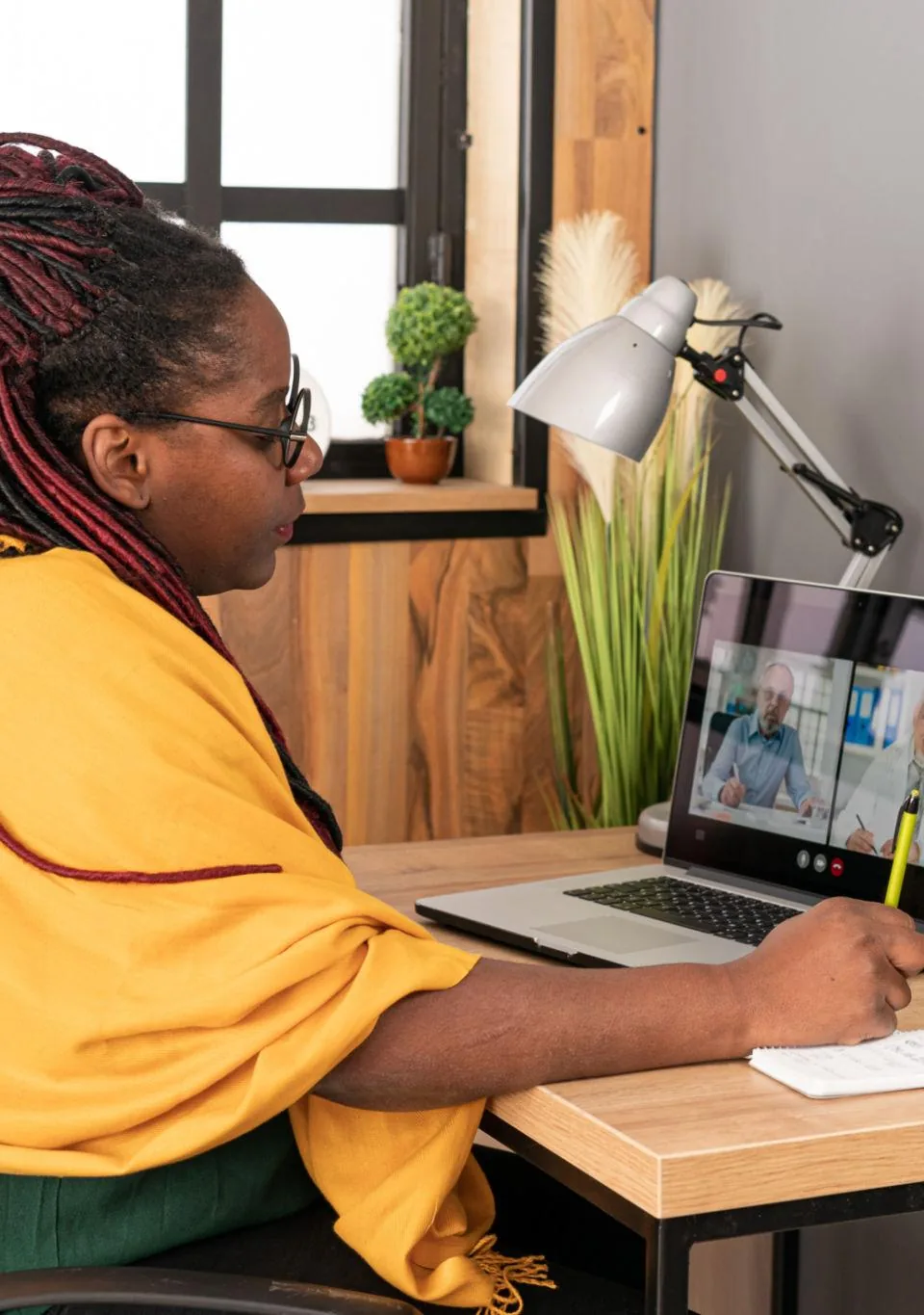 A woman taking notes at a home desk while a video call runs on her laptop